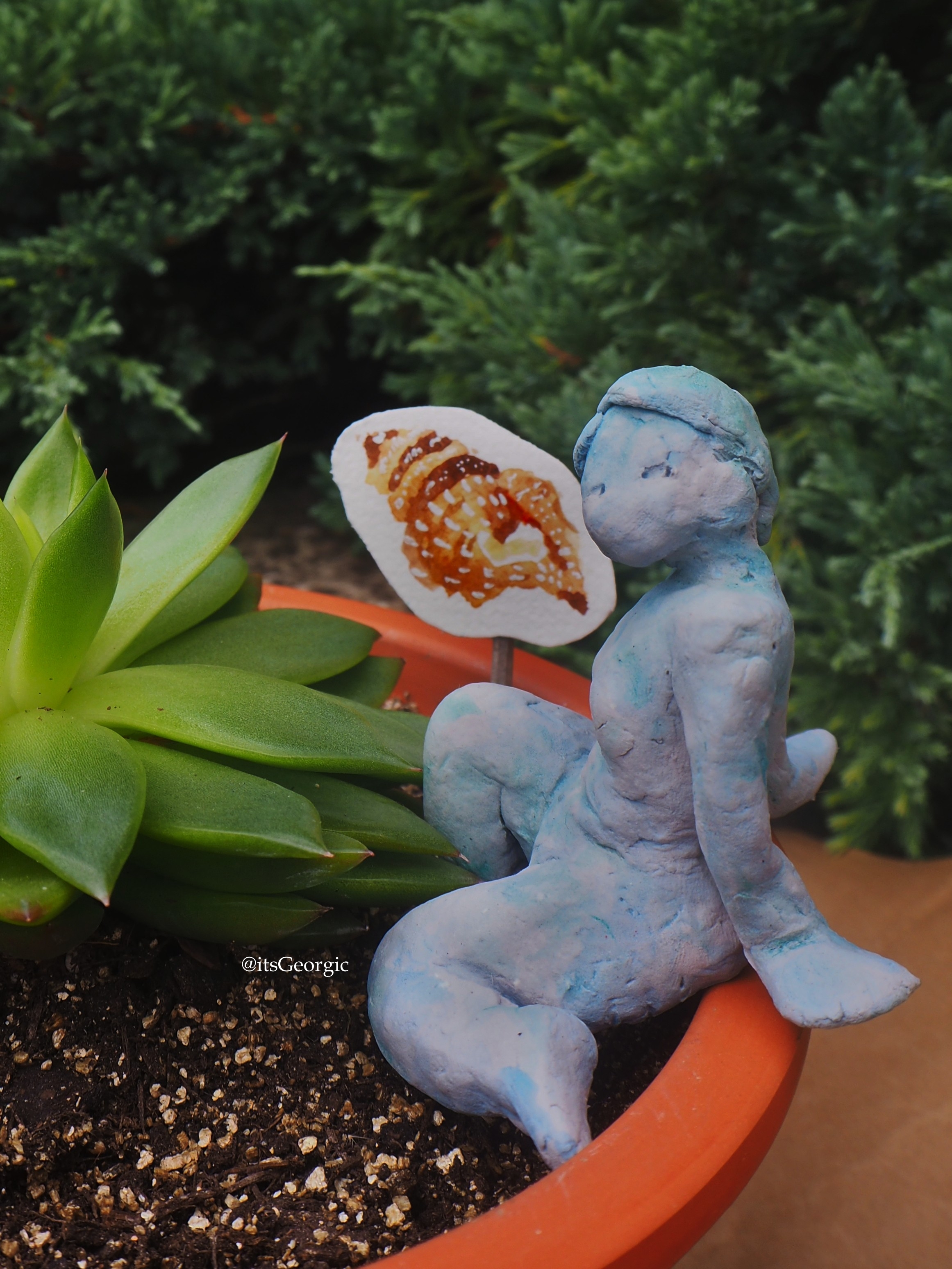 Photo de la statuette Bolina posée sur le bord d'un pot contenant une succulente (echeveria agavoides) et devant un dessin à l'aquarelle d'un coquillage plantée dans la terre