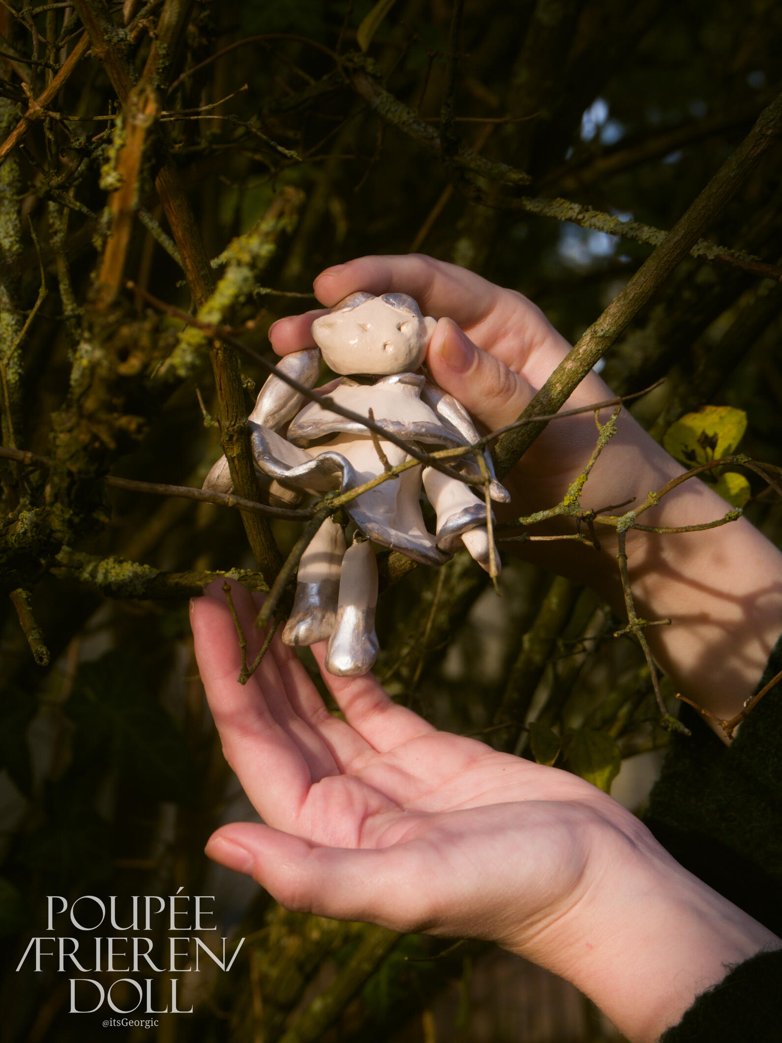 Photo de la poupée articulée dans un arbre, assise sur une branche les jambes ballantes. Des mains humaines l'encadrent.