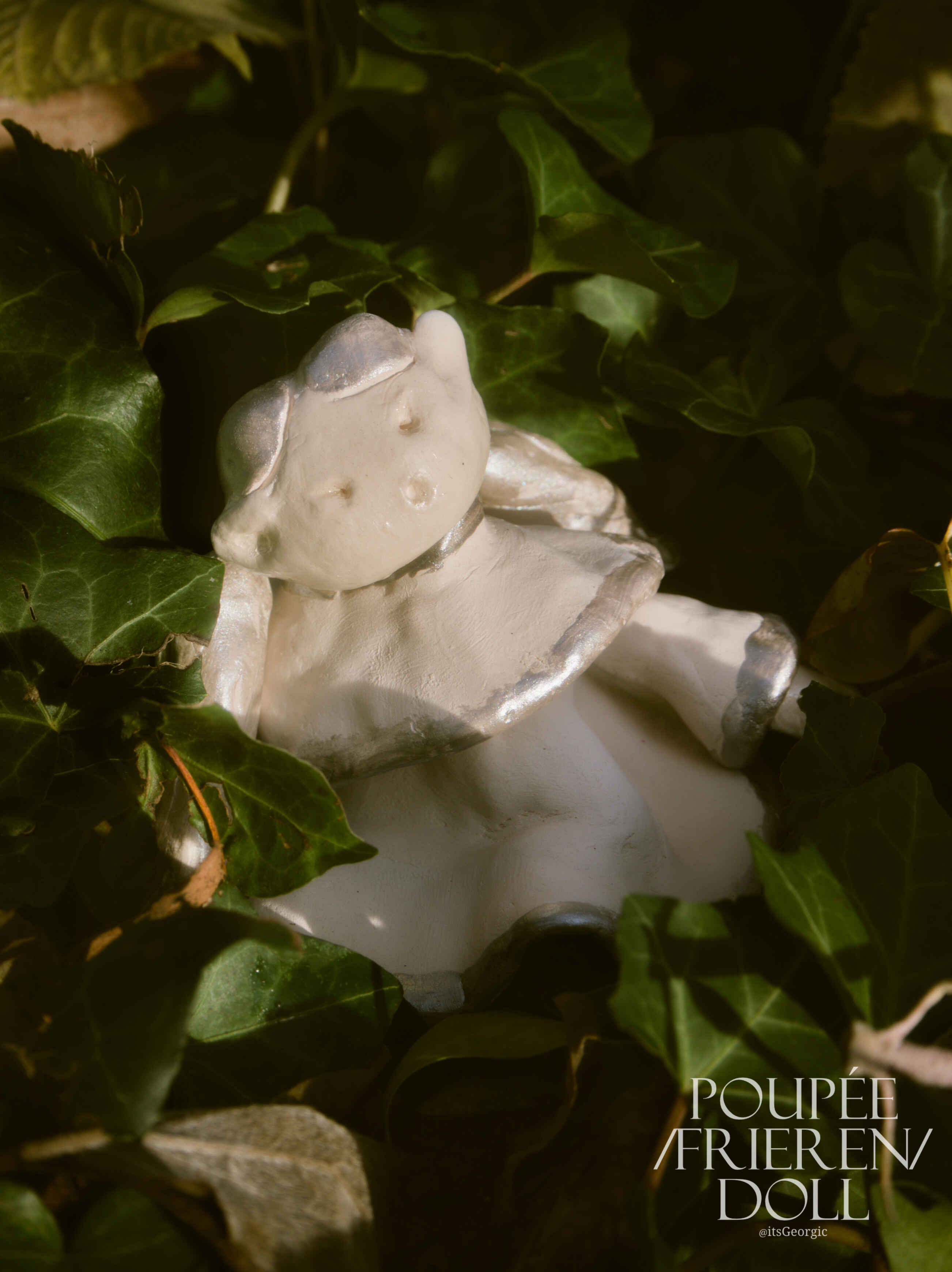 Photo d'une poupée inspirée de l'anime Frieren. La poupée est en céramique avec des accents argentés et posée dans un pare-terre de lierre.