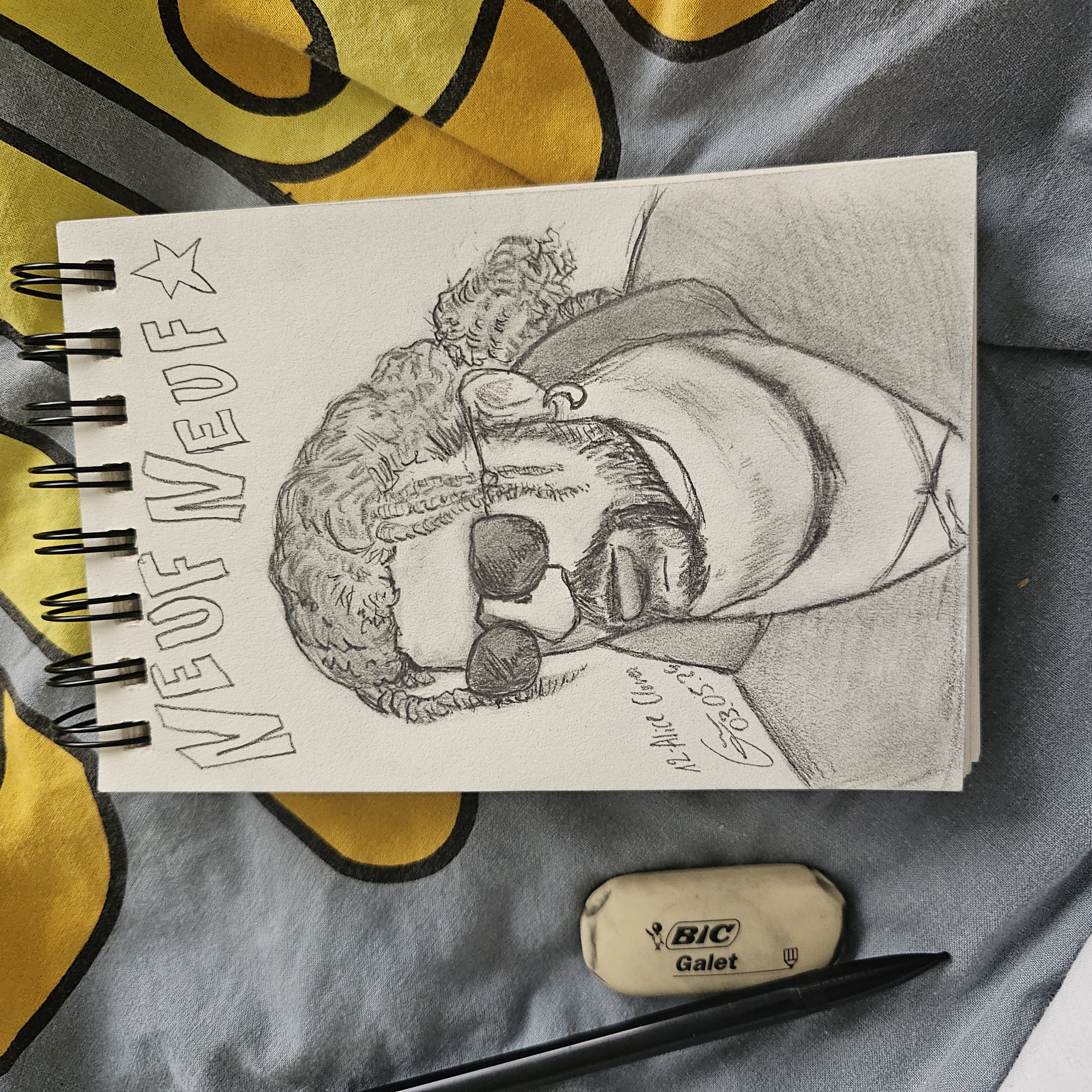 Photo d'un portrait au crayon dans un carnet de croquis. L'homme a des lunettes de soleil et des locs dans les cheveux. Il porte un col roulé et a une boucle d'oreille de lune